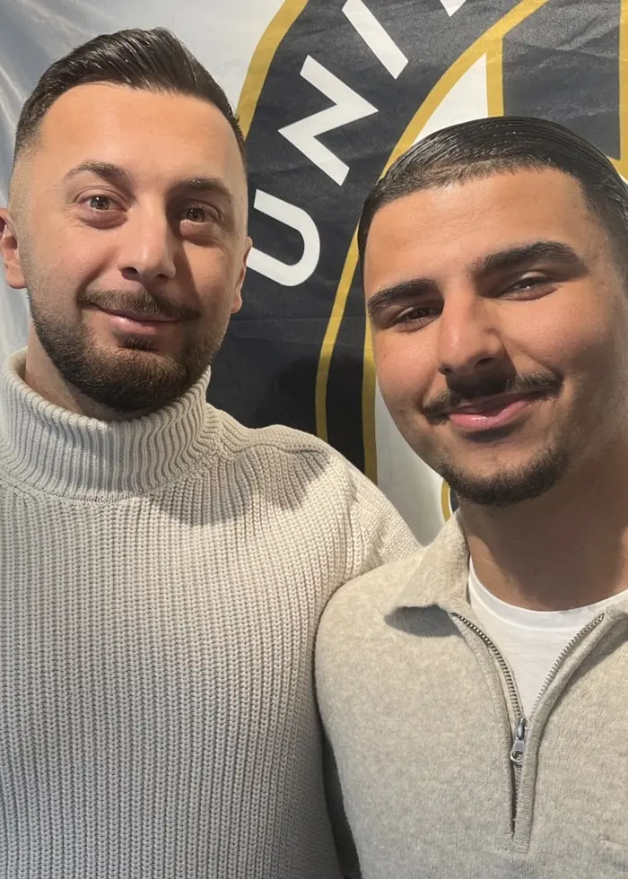 Två personer står nära varandra framför en vägg med en stor logotyp för United People. De bär ljusa tröjor och i bakgrunden syns upphängda kläder.