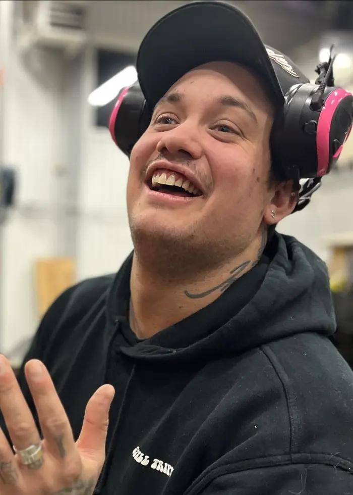 En person med hörselskydd arbetar vid en trävägg där diverse verktyg och utrustning är monterade. Bakgrunden visar en lagerlokal med hyllor och kartonger.