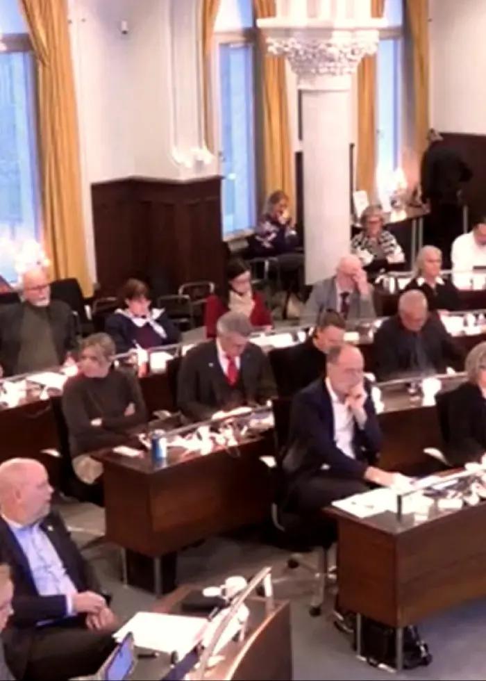 Bild från en regionfullmäktigesal med flera personer sittande vid bord. Salen är inredd med höga fönster och gula gardiner.