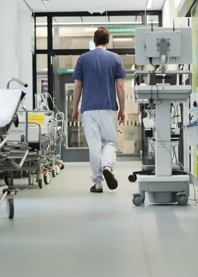 En person i sjukvårdskläder går genom en ljus korridor på en akutmottagning. Sängar och medicinsk utrustning står längs väggarna. Bilden är tagen på Danderyds sjukhus i Stockholm.