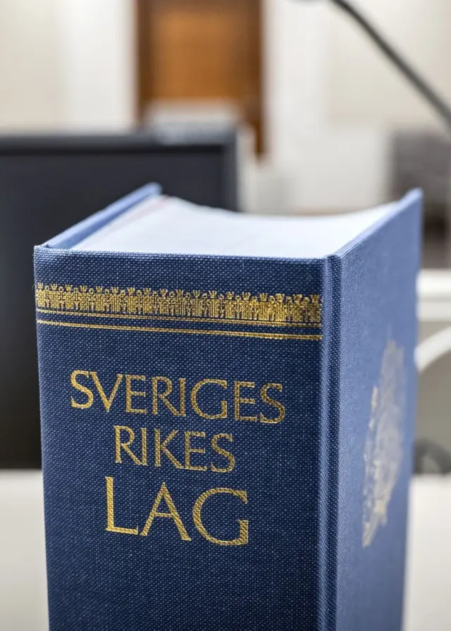 En närbild av en blå bok med texten 'Sveriges rikes lag' placerad på ett bord i en rättssal. I bakgrunden syns en datorskärm, mikrofon och möbler i en modern domstolsmiljö.