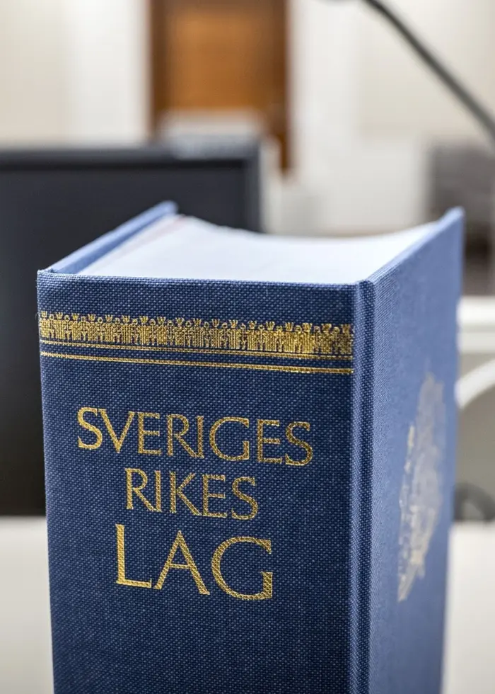 En närbild av en blå bok med texten 'Sveriges rikes lag' placerad på ett bord i en rättssal. I bakgrunden syns en datorskärm, mikrofon och möbler i en modern domstolsmiljö.