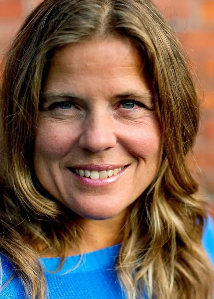 En kvinna med långt brunt hår klädd i en blå tröja står framför en tegelvägg. Bilden är tagen i ett stadsmiljö med fokus på personen.