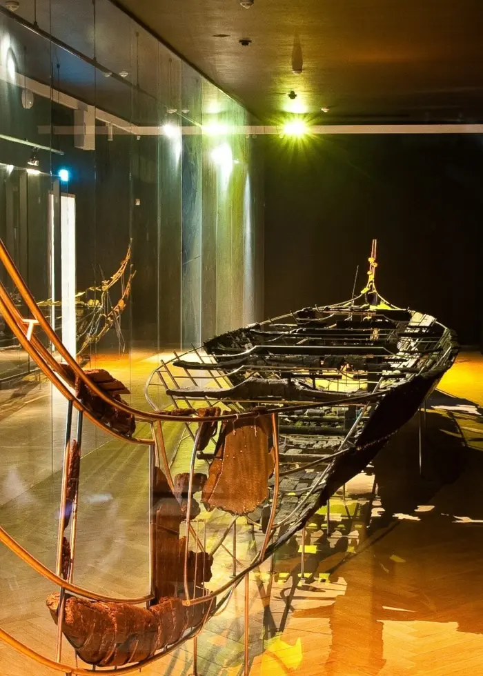 En utställning av Hjortspringsbåten, en rekonstruerad gammal träbåt från nordisk järnålder, belyst med spotlights och omgiven av mörka draperier. Båten är placerad i en modern museimiljö.