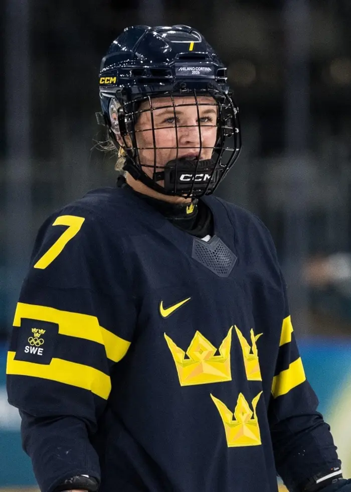 Två personer kopplade till ishockey ses i bilden. Den ena är en spelare i mörkblå landslagsdräkt med tre kronor på bröstet. Den andra bär en mörk jacka och mössa med svenska flaggan, och gör en tumme upp.
