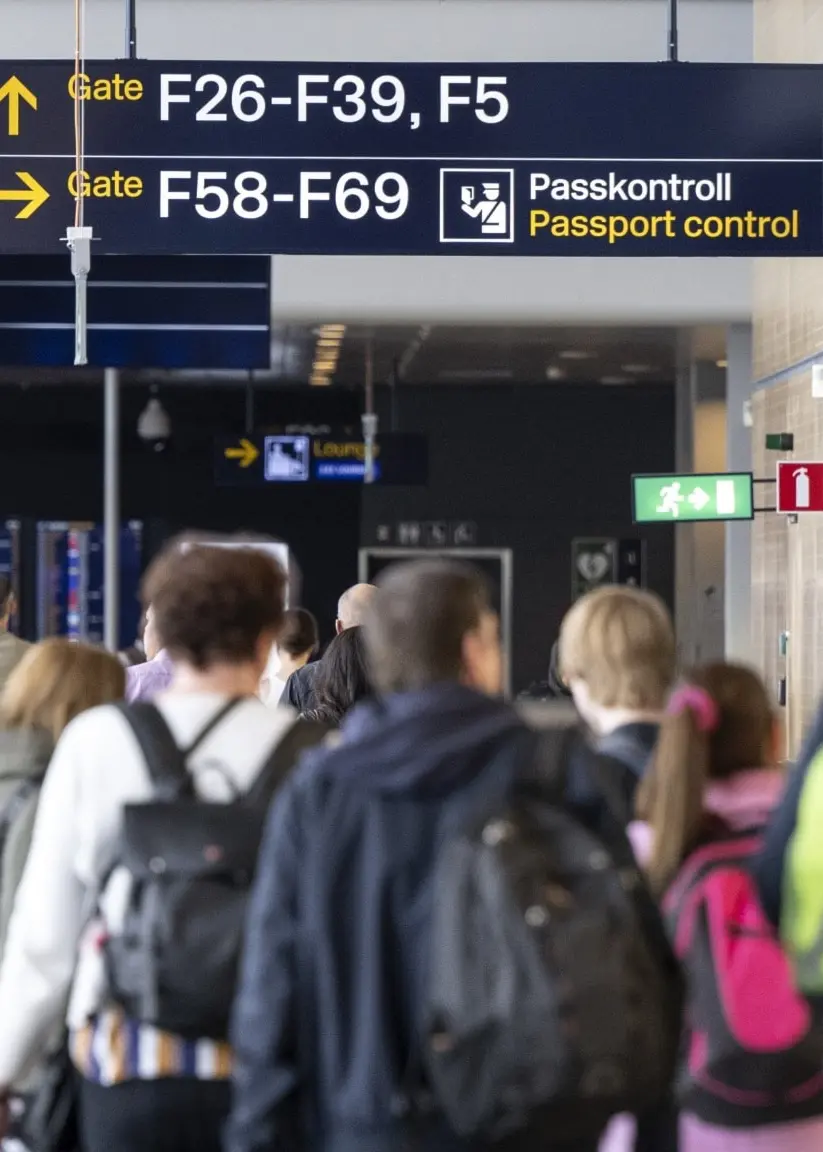 Passagerare med ryggsäckar och bagage går genom en terminal på en flygplats. Skyltar visar vägen till olika gateområden.