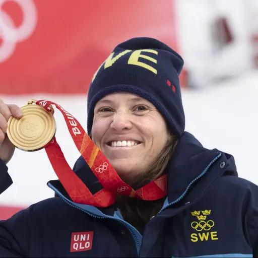 En person klädd i svenska landslagets vinterkläder håller upp en guldmedalj framför en röd bakgrund med OS-ringarna synliga. Bilden är från vinter-OS 2022 vid alpina arenan i Yanqing.