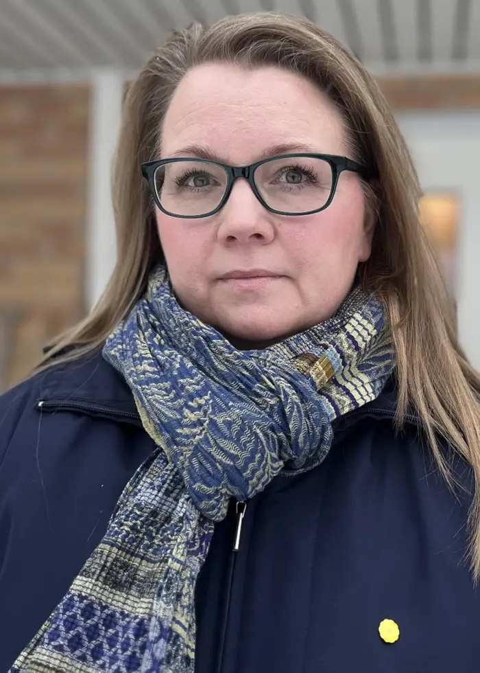 Sara Brodin står utomhus framför en byggnad med vita dörrar och tegelvägg. Hon bär en blå jacka och en mönstrad halsduk.