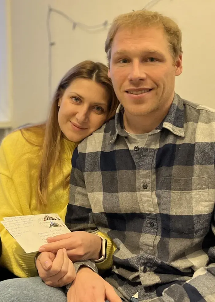 Två personer sitter i en soffa i ett vardagsrum. En person håller ett kort i handen. I bakgrunden syns ett fönster med gardiner, en krukväxt och en byrå.