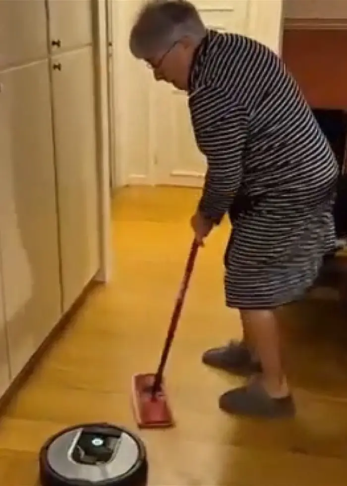 En kvinna i randig morgonrock och tofflor står i ett kök och låtsas curla med en robotdammsugare. Hon håller en sopborste framför sig och ser ut att ha roligt.