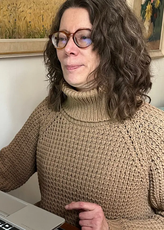 En person sitter vid ett skrivbord och arbetar på en bärbar dator i ett hemlikt rum med tavlor på väggen och en soffa i bakgrunden. På bordet ligger en anteckningsbok och en penna.