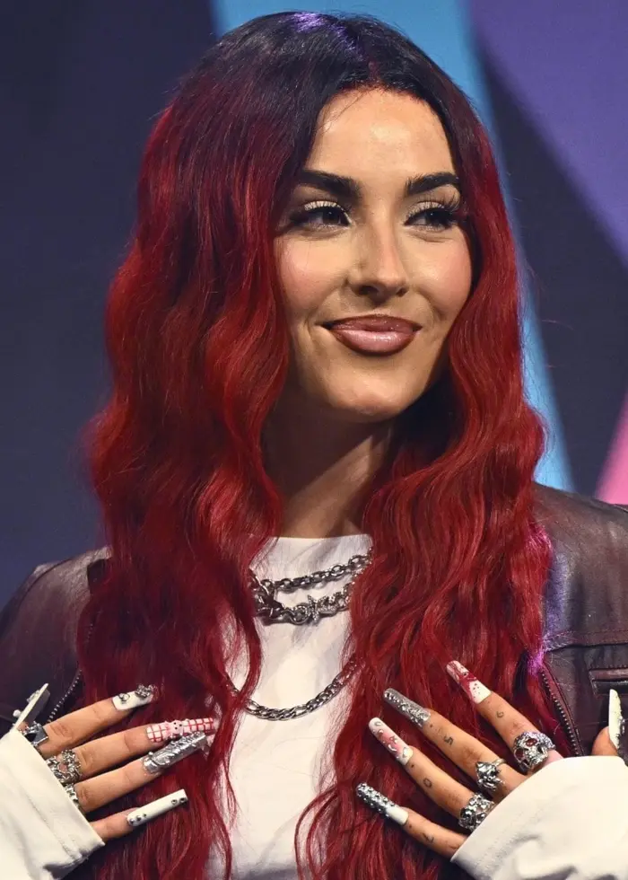 En artist med långt rött hår står på en scen med färgglada geometriska former i bakgrunden. Hon är klädd i en vit topp, en mörk jacka och har händerna vid brösthöjd.