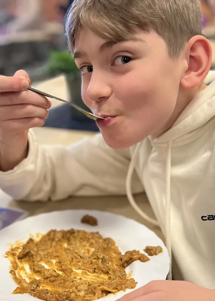 En elev sitter vid ett bord i en skolmatsal och äter mat från en vit tallrik. På bordet finns även en meny och andra elever i bakgrunden. Personen bär en ljus hoodie.