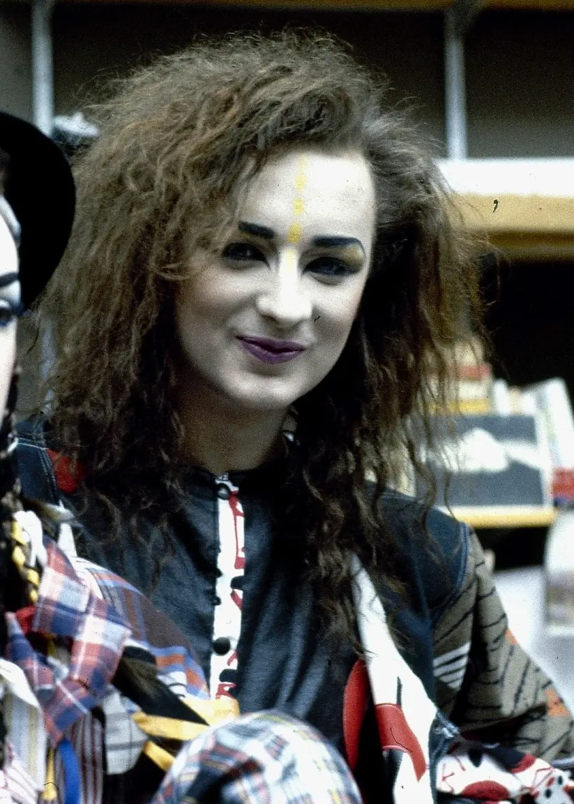 Boy George i Culture Club står jämte sig själv som vaxdocka. Han håller om vaxdockan med sin högra arm. Han ser glad ut. Han har långt lockigt hår.