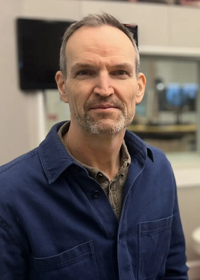 En person står i en radiostudio med mikrofoner och studioutrustning i förgrunden. I bakgrunden syns en glasvägg som separerar studion från ett annat rum.