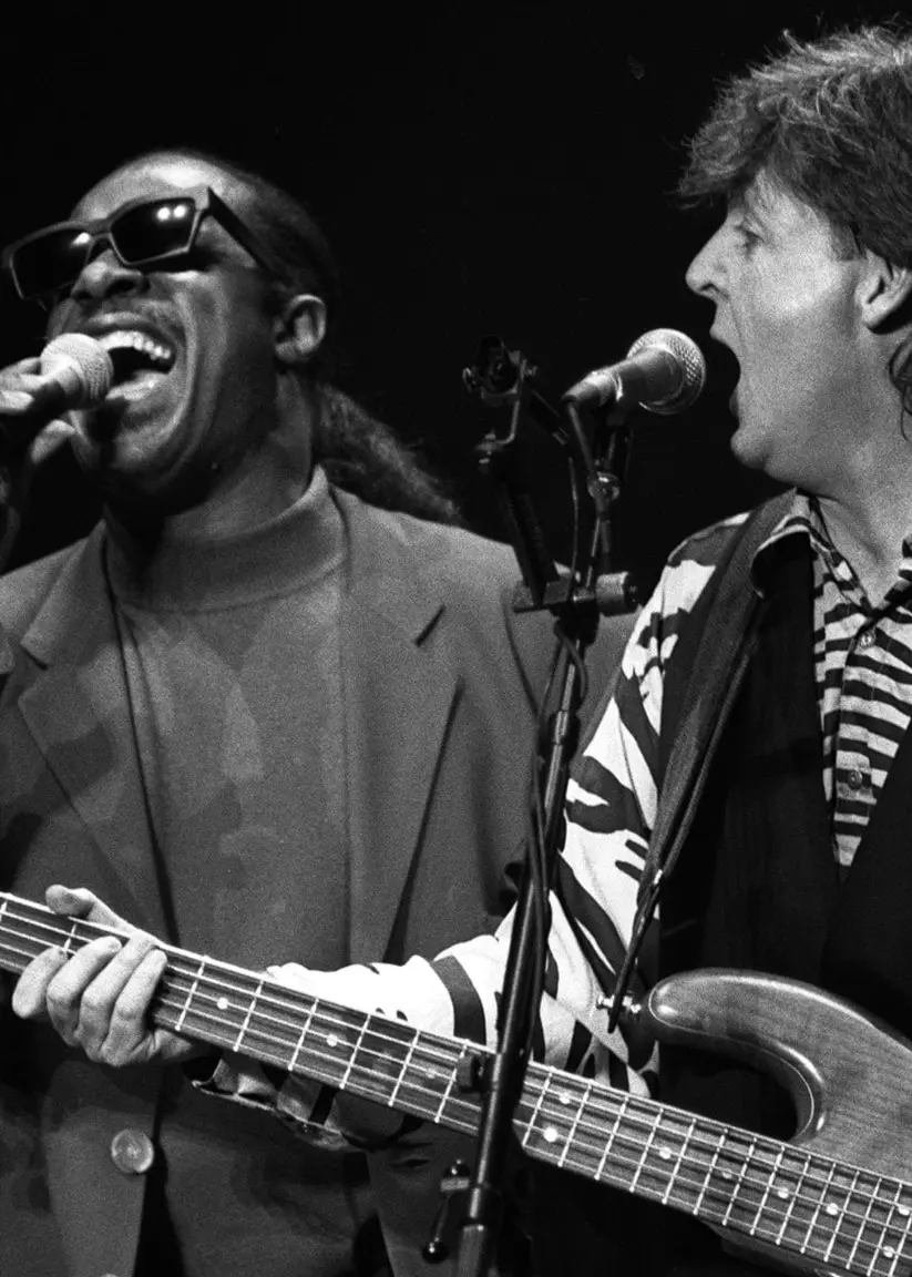 Stevie Wonder står till vänster och sjunger i en mikrofon som han håller nära munnen med höger hand. Jämte honom står Paul McCartney och sjunger i en mikrofon som står på ett stativ. Han håller en gitarr och spelar på.