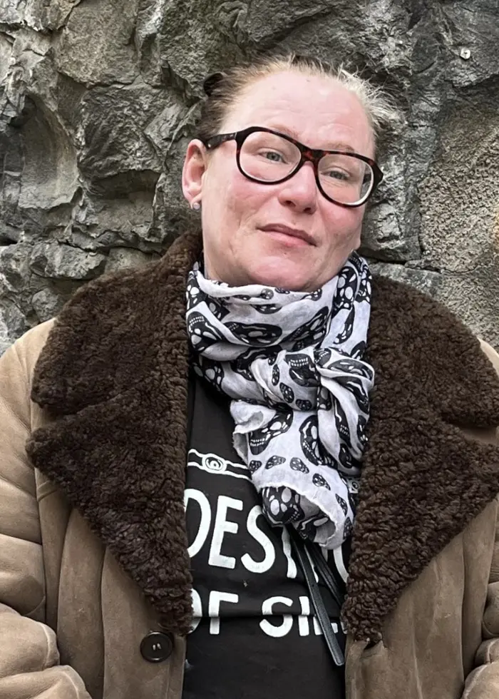 Marilyn Winter i beige kappa står framför en stenmur med graffiti i bakgrunden. Personen håller i en tidning och är klädd i mörka kläder under kappan.