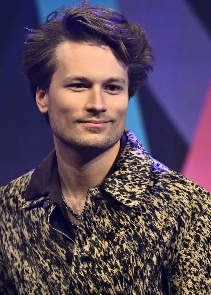 En person med lockigt hår står på en scen med en färgglad bakgrund i form av diagonala mönster i rosa, blått och svart. Personen bär en mönstrad skjorta och deltar i en presentation.