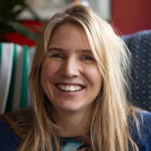 Amanda Romare sitter i en fåtölj med en blå filt bakom sig. Bakgrunden visar färgglada ljusslingor och väggdekorationer i ett hemtrevligt rum.