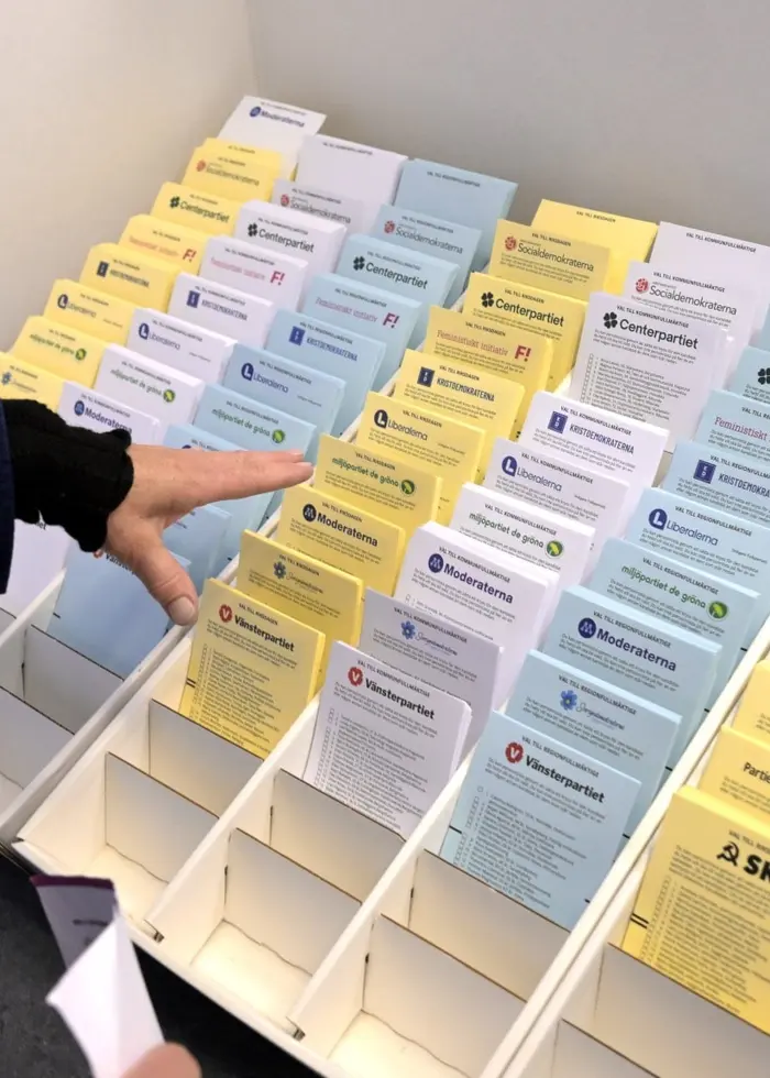 En person pekar på ett ställ med valsedlar i olika färger vid ett röstningslokal under ett val i Sverige.