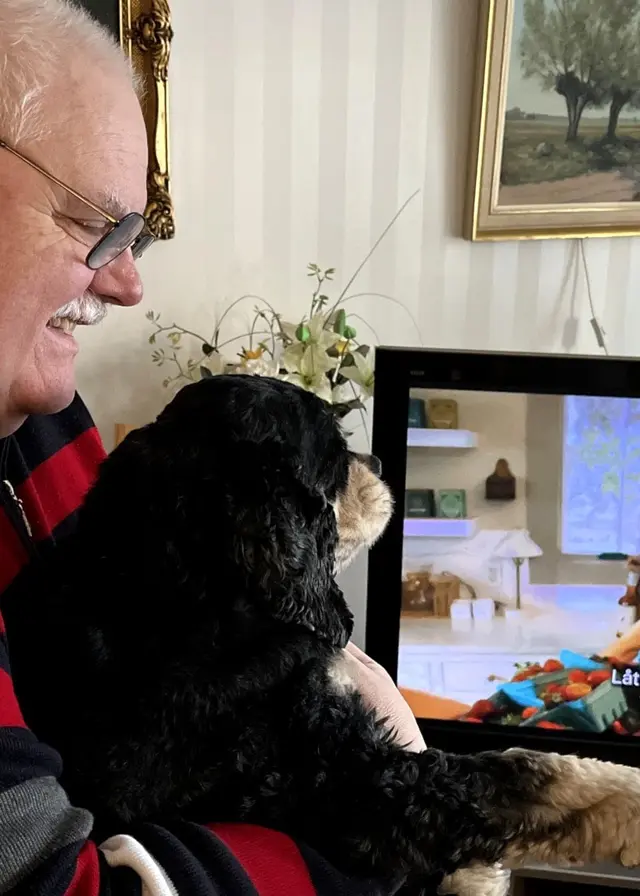 Husse Karl-Lennart Säll håller i sin hund framför en tv. Karl-Lennart ler mot hunden Pumba som har fullt fokus på tv:n.