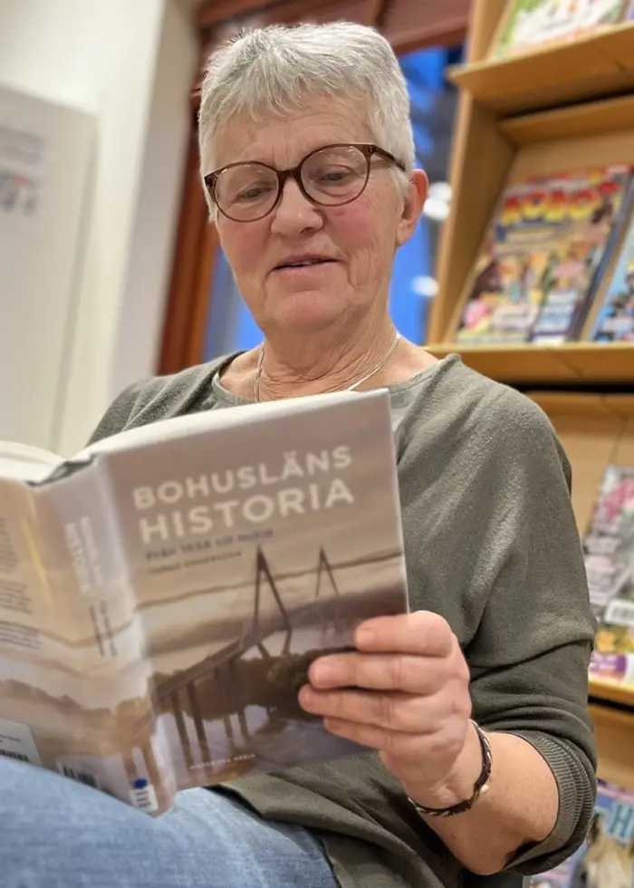 En person sitter i ett bibliotek och läser en bok med titeln 'Försvunnen historia'. I bakgrunden syns bokhyllor fyllda med tidskrifter och böcker. En svart ryggsäck står på golvet bredvid personen.