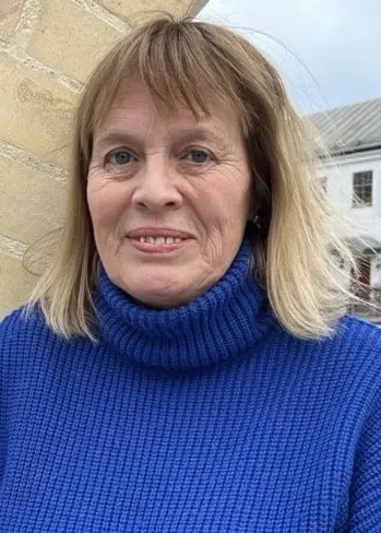 Mariette Lindstein står framför en tegelvägg dekorerad med blå mosaik, iklädd en blå stickad tröja. I bakgrunden syns en stadsmiljö med byggnader och trottoar.