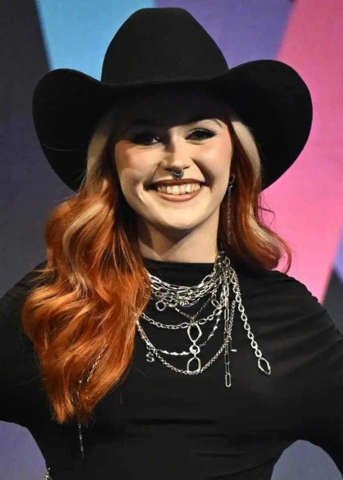 Eva Jumatate står på en scen under en presskonferens i Stockholm inför Melodifestivalen 2026. Hon är klädd i en svart hatt, svart klänning och flera halsband och står framför en färgglad bakgrund med geometriska mönster.