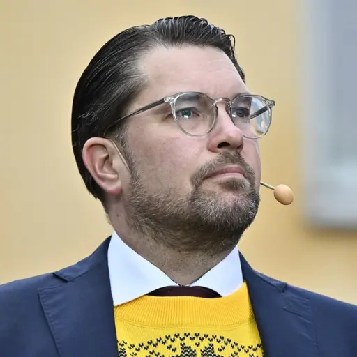 En man i blå kostym och gul tröja med mönster står utomhus framför en gul byggnad. Han har en mikrofon vid kinden och kort, mörkt bakåtkammat hår.