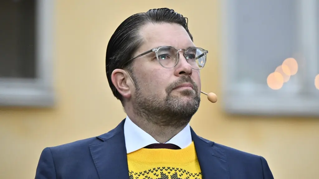 En man i blå kostym och gul tröja med mönster står utomhus framför en gul byggnad. Han har en mikrofon vid kinden och kort, mörkt bakåtkammat hår.