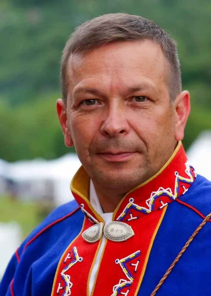 En person klädd i traditionell samisk dräkt med blå och röda färger samt dekorativa detaljer står utomhus med grön natur och vita tält i bakgrunden.