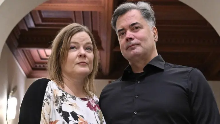 Två personer står i en elegant byggnad med valv och högt i tak. Mannen bär mörka kläder, och kvinnan en mönstrad blus. De verkar vara i en formell miljö, möjligtvis inför en rättslig process.