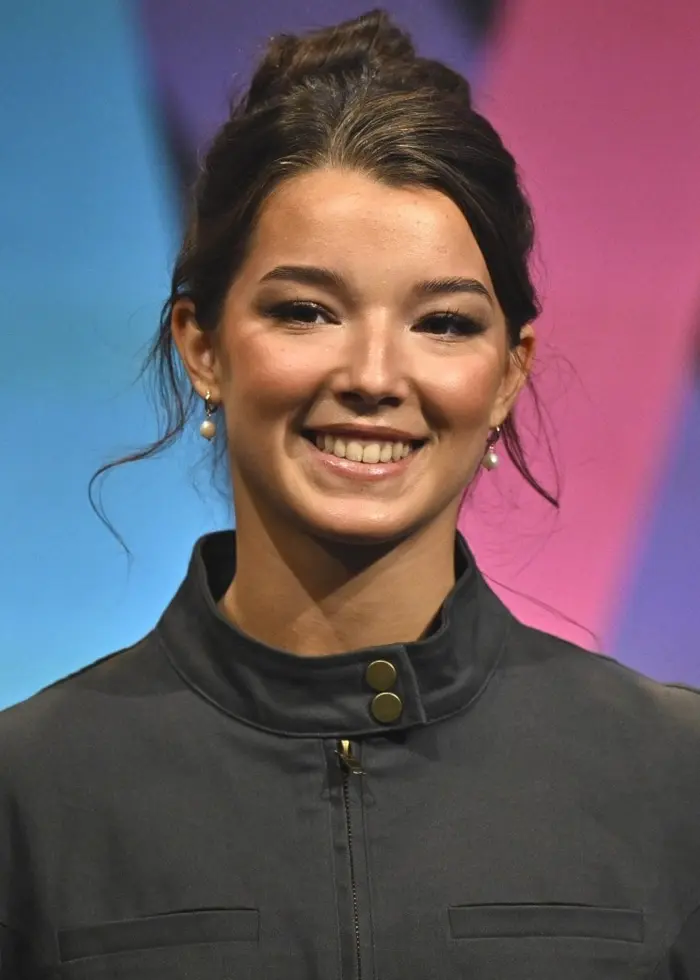 Eva Jumatate står på en scen under en presskonferens i Stockholm inför Melodifestivalen 2026. Hon är klädd i en grå skjorta och står framför en färgglad bakgrund med geometriska mönster.