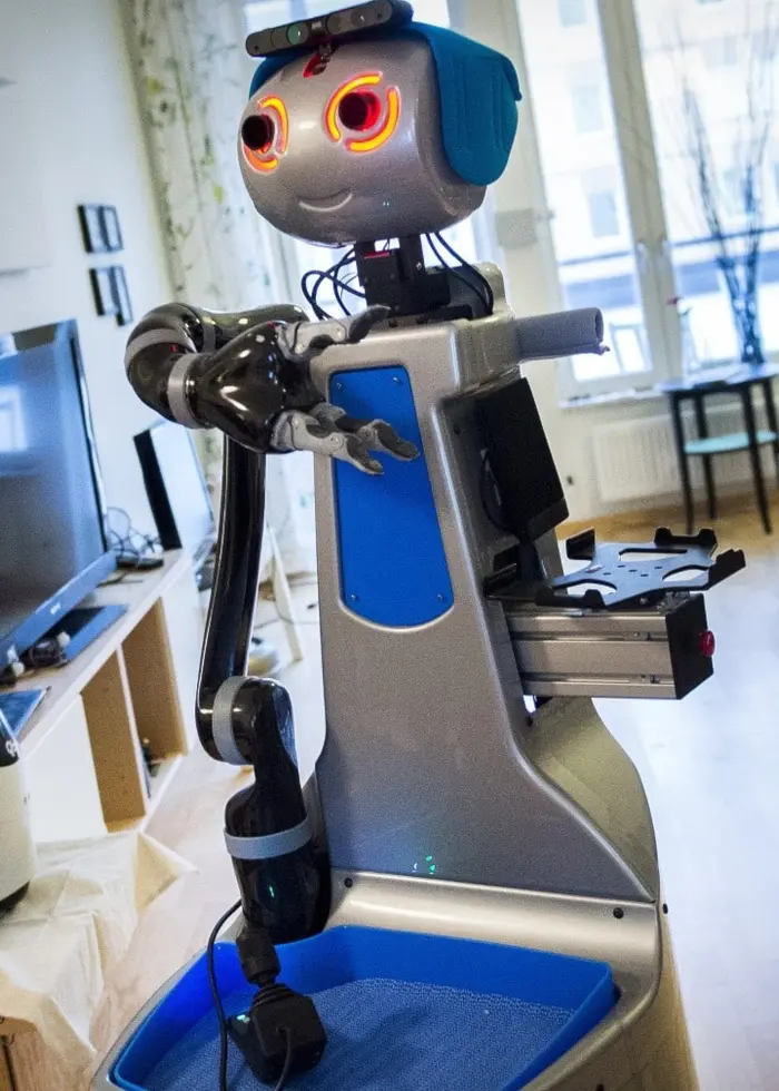 En robot rör sig genom ett modernt vardagsrum för att hämta medicin till en äldre kvinna som sitter i en soffa.