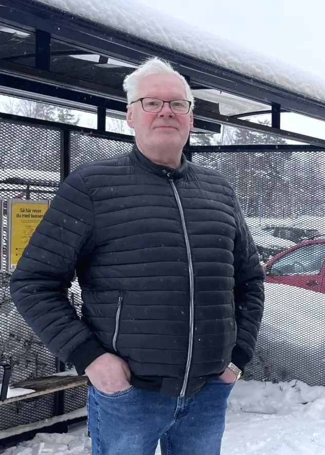 Två personer står framför en gul buss med numret 801 tydligt synligt på displayen. Bussens hållplatsområde är täckt av snö, och bakgrunden visar parkerade bilar och ett staket.