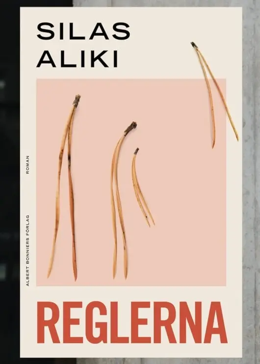 En person står framför en betongvägg, klädd i en svart läderliknande kortärmad skjorta över en vit tröja. Till vänster syns en bok med titeln 'Reglerna' av Silas Aliki.