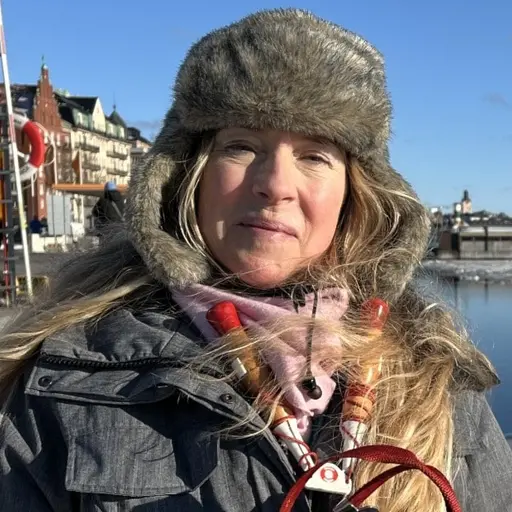 En person står utomhus vid en frusen vattenyta med byggnader i bakgrunden. Personen håller ett husdjur i famnen och är klädd i vinterkläder. Solen skiner och himlen är klarblå.