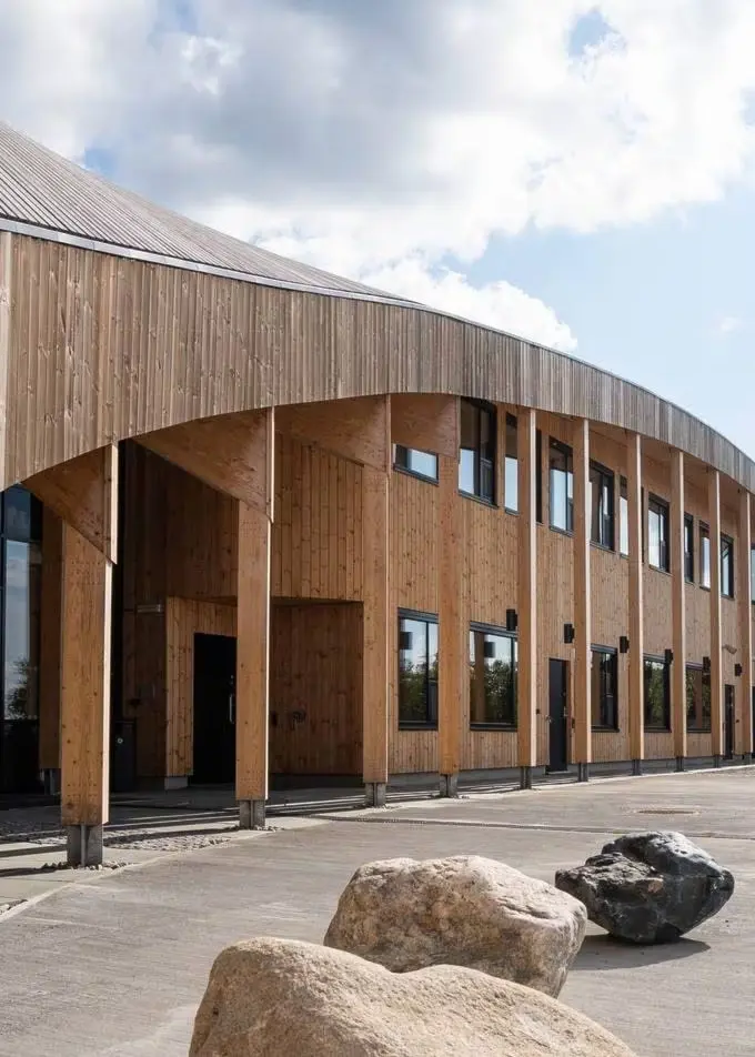 Byggnad med modern arkitektur i trämaterial, belägen i naturskön miljö med stenformationer på marken och en klar himmel med moln i bakgrunden. Platsen är Čoarvemátta i Kautokeino.
