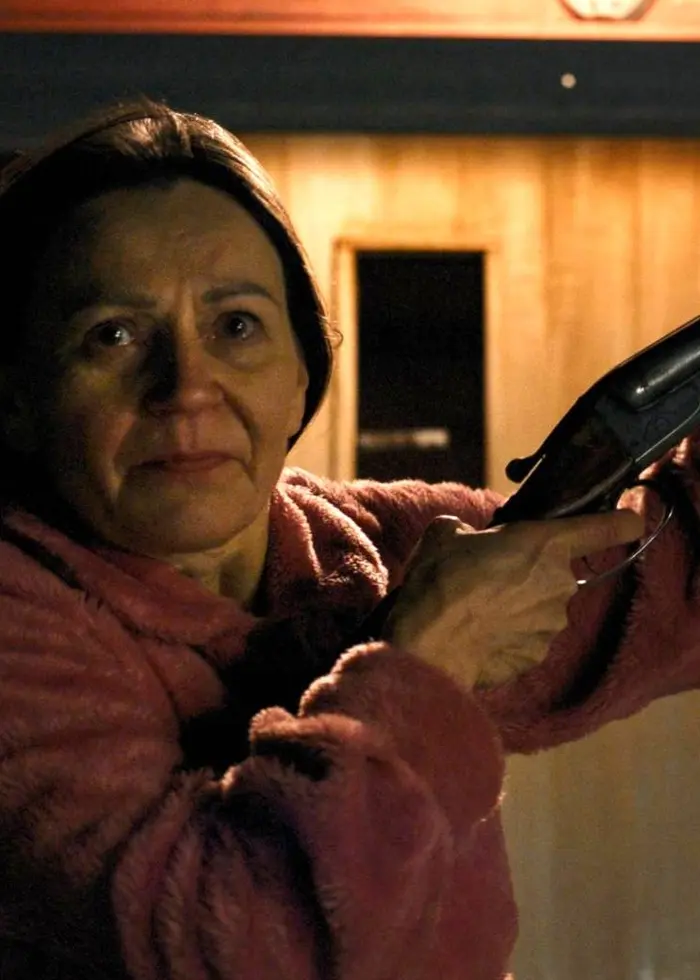 En person håller upp ett gevär utanför en träbyggnad, klädd i en rosa tröja. Miljön är mörk med belysning från en lampa ovanför dörren. Scenen har en dramatisk atmosfär.