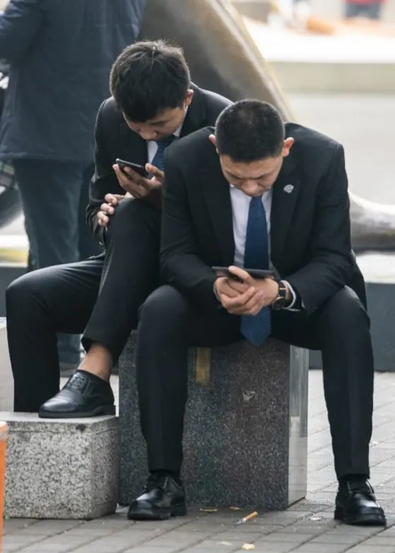 Bild från Peking, Kina, med flera affärsmän klädda i kostym som sitter på en bänk och tittar på sina mobiltelefoner. I bakgrunden syns en stor skulptur och människor som promenerar i stadsmiljön.