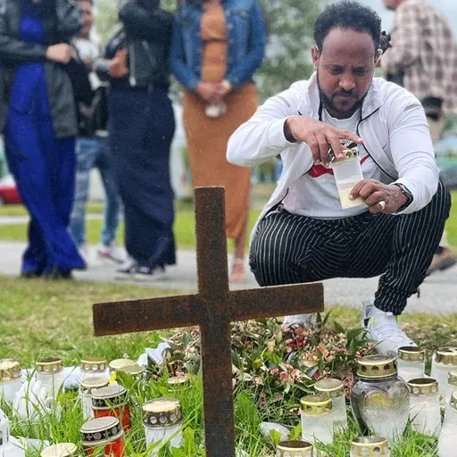 Två personer tänder ljus vid en minnesplats med ljuslyktor, blommor och ett kors. I bakgrunden står flera personer.