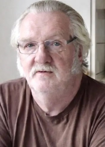 En man med brun tröja, grått hår och glasögon som sitter i ett kök.