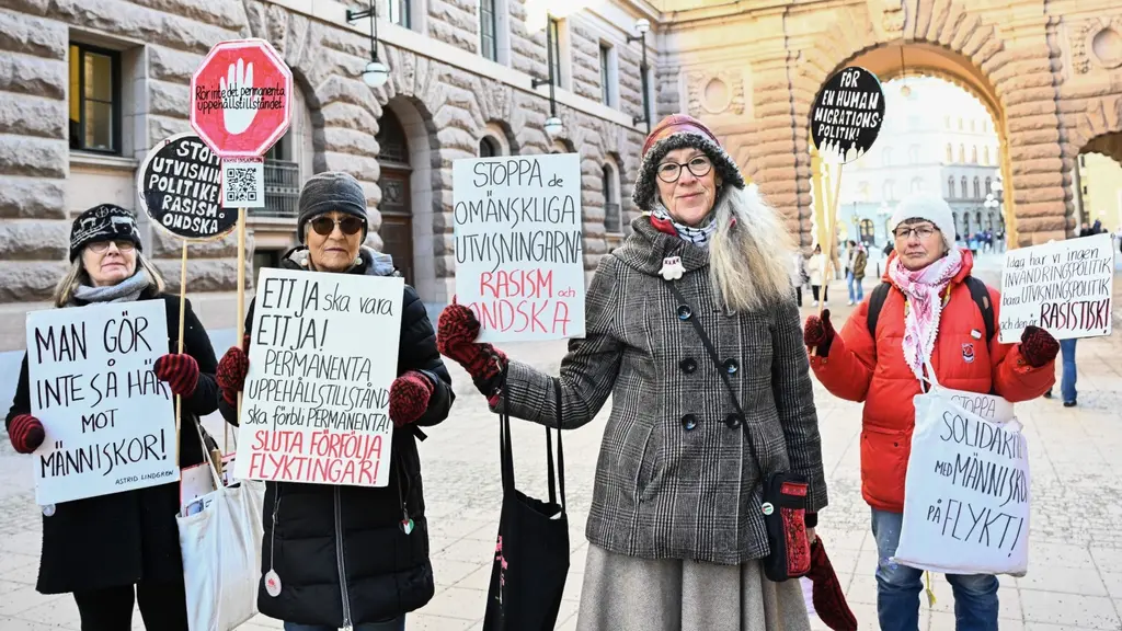 Fyra personer står utanför Sveriges riksdag med skyltar som protesterar mot utvisningar. De är varmt klädda och håller upp budskap som uppmanar till medmänsklighet. Solen lyser på platsen.
