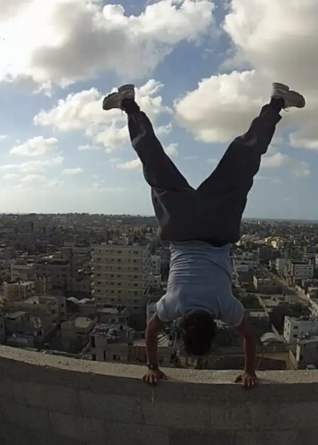 En person utför en handstående på en hög mur med en stadsvy i bakgrunden under en klar himmel med moln. Bilden förmedlar en känsla av frihet och äventyr.