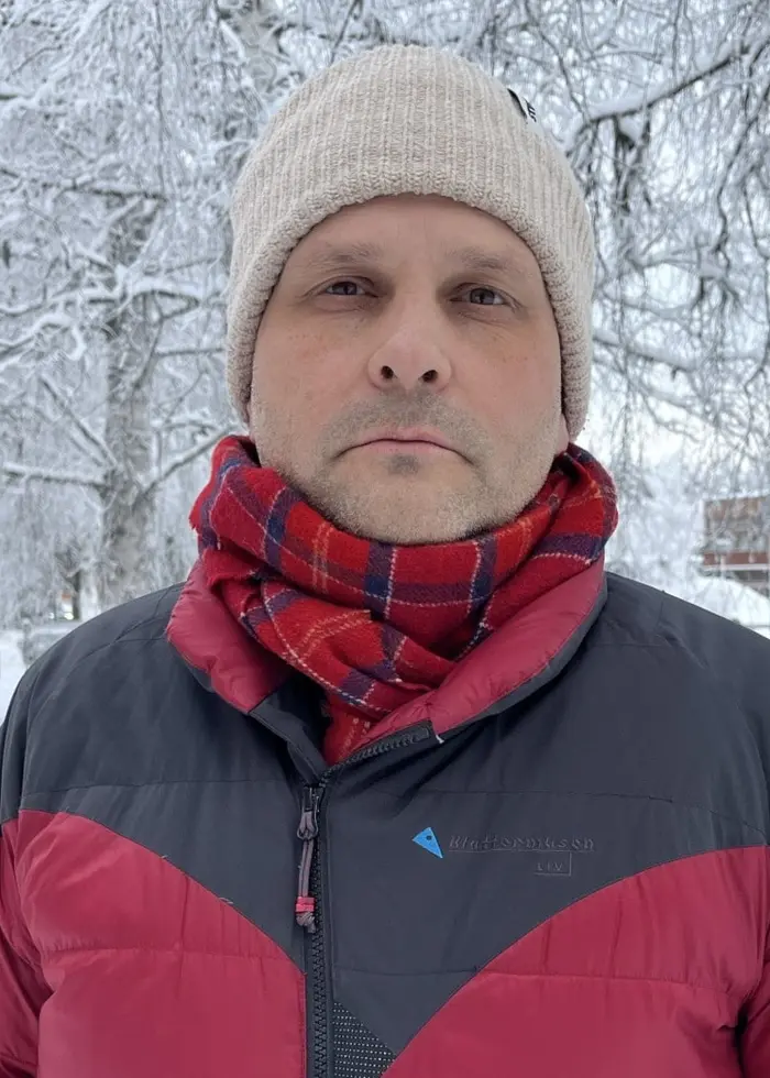 En person klädd i röd vinterjacka och beige mössa står utomhus i en snöig miljö med frostiga träd och parkerade bilar i bakgrunden.