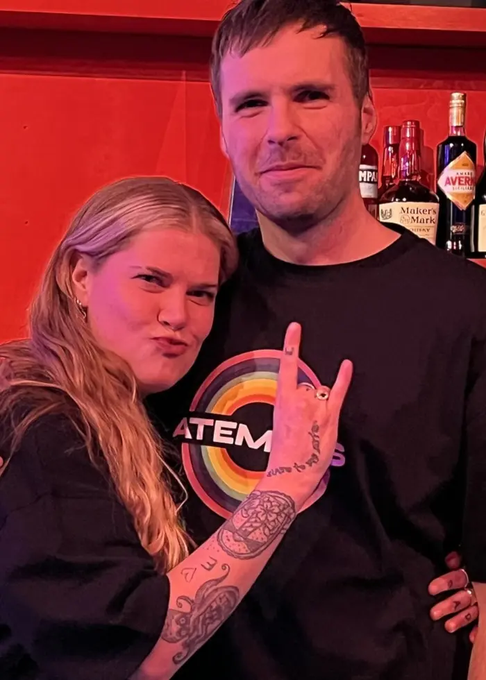 Två personer iklädda svarta tröjor står framför en bardisk med röda väggar och hyllor fyllda med flaskor. En av personerna gör en rockgest med handen.