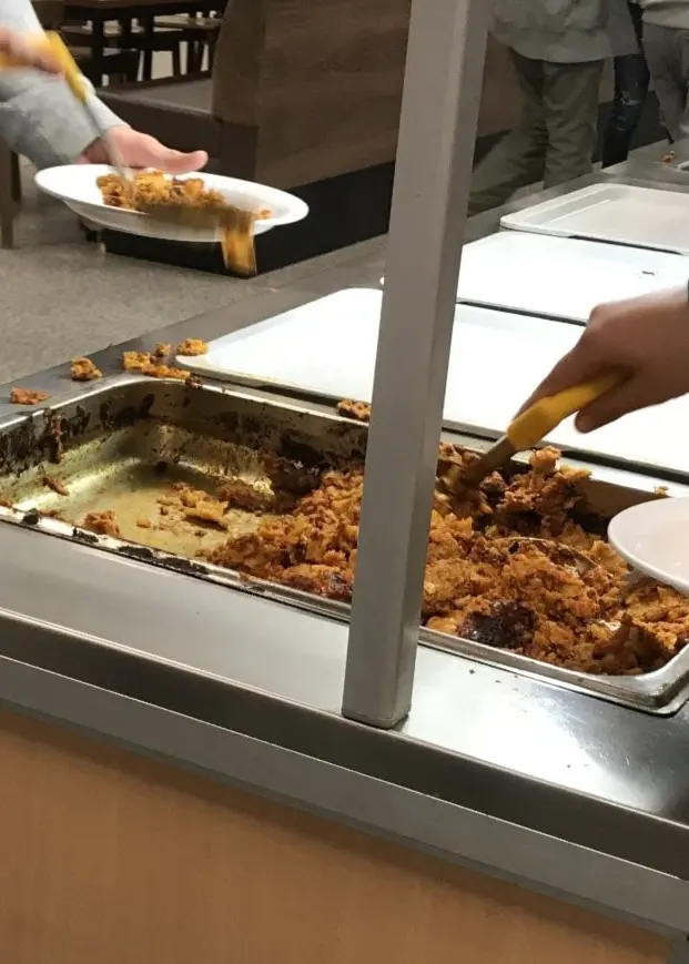 Ett vildsvin springer på en gräsyta till vänster, och till höger syns en skolmatsal där mat serveras från en buffé.