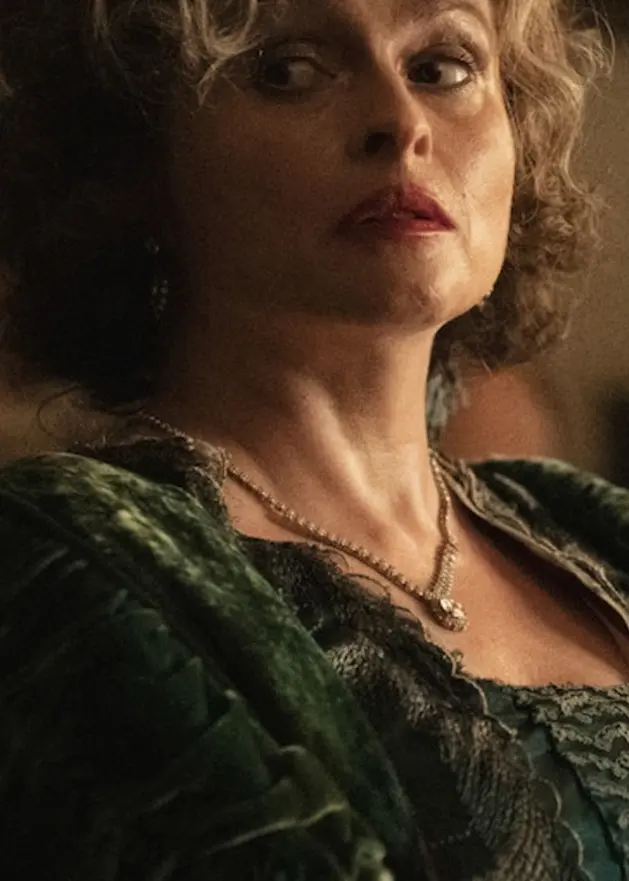 Två personer i historiska kläder befinner sig i ett dunkelt och elegant rum. Den ena bär en grön klänning med detaljerad struktur, medan den andra håller en mask och är klädd i ljusa färger.