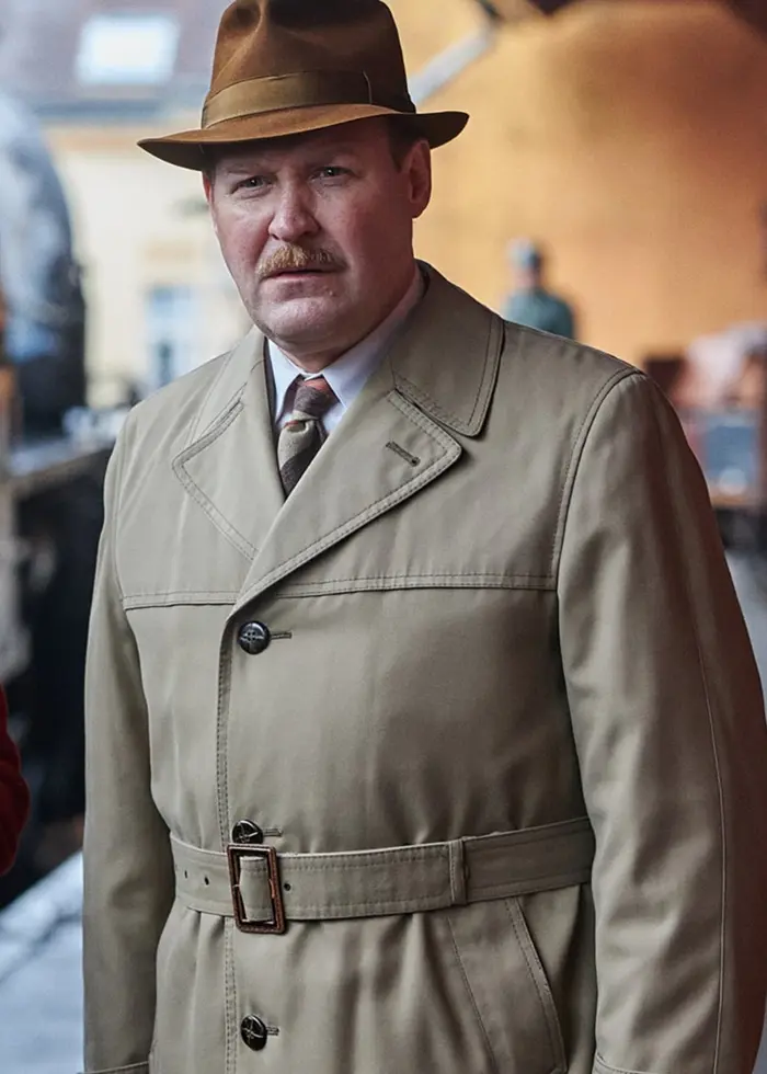 Två personer i tidstypiska kläder från mitten av 1900-talet står på en tågperrong. En av dem håller i en kamera och bär röd kappa och grön mössa, medan den andra har trenchcoat och hatt.