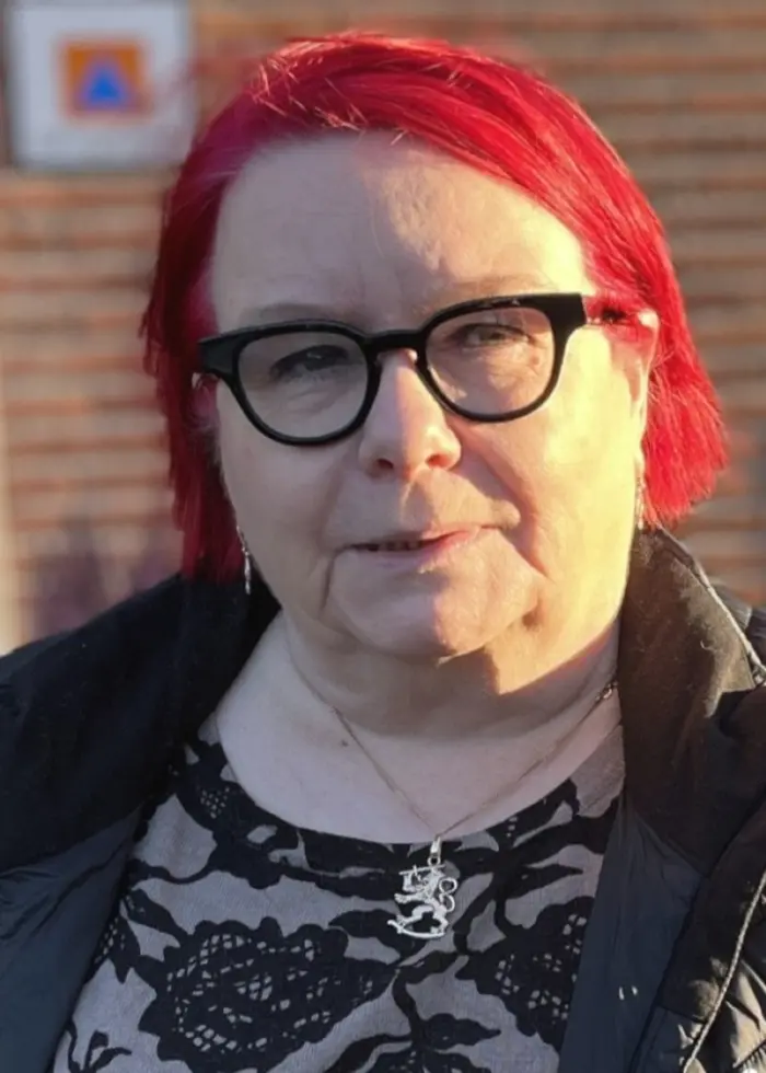 En kvinna med röd hårfärg står utomhus framför en tegelvägg. Hennes ansikte är suddat. På bilden syns symboler som representerar olika minoriteter, inklusive en romsk flagga och en Davidsstjärna.